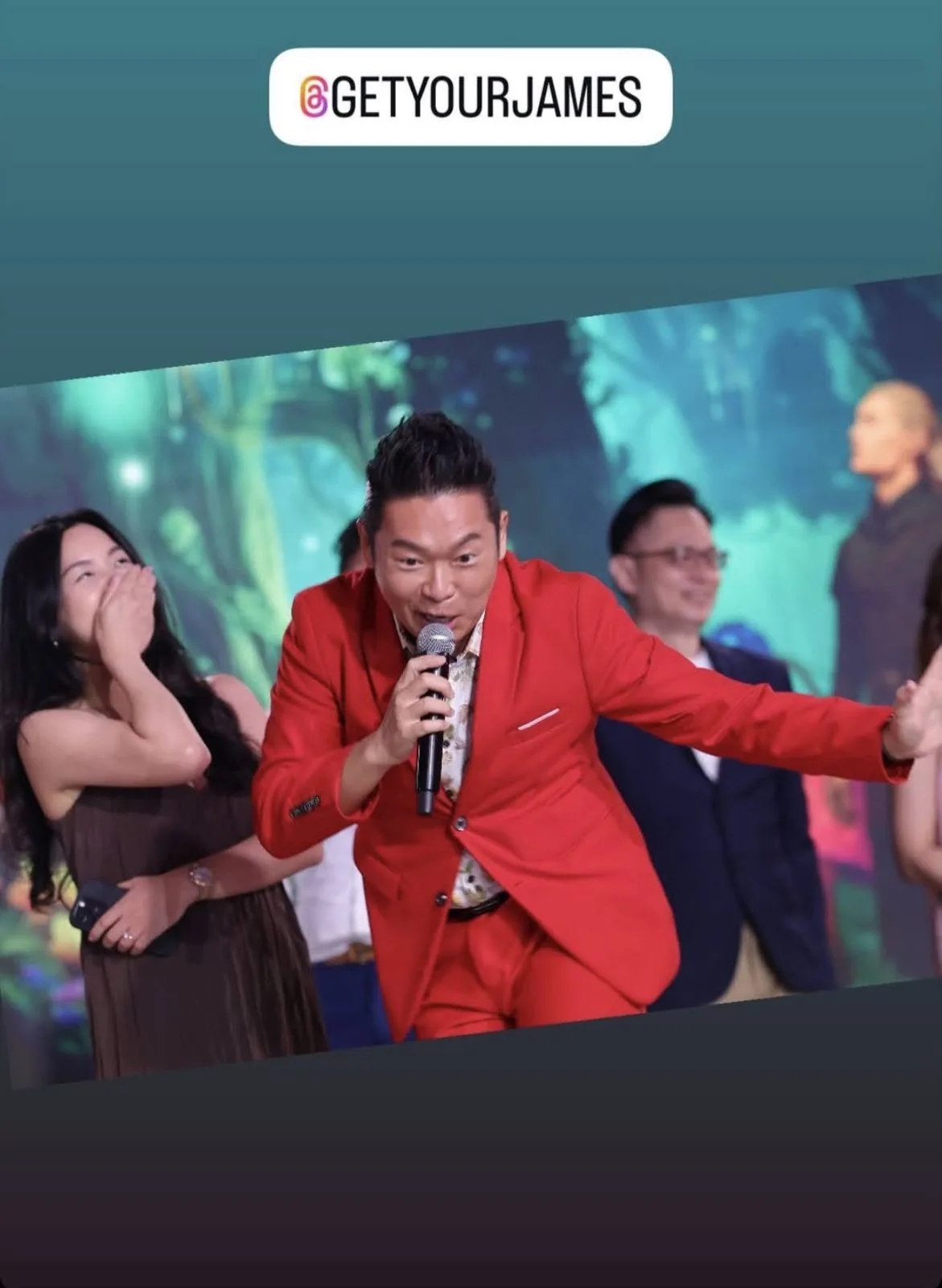 James Yang in a red suit hosting on stage, guests laughing behind him